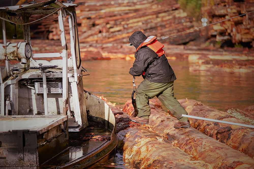 A man wearing the Valdor 8" Waterproof Comp Toe Insulated Logger boots in brown and carrying a fishing pole and walking along logs to get to their boat.